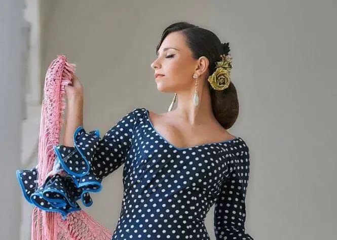 Mujer con vestido azul marino de lunares sosteniendo un chal rosa estampado sobre un fondo gris.
