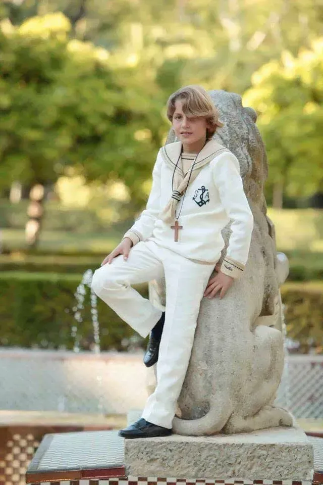 Una persona vestida de blanco posa sobre una estatua de un león de piedra junto a una fuente en un parque.