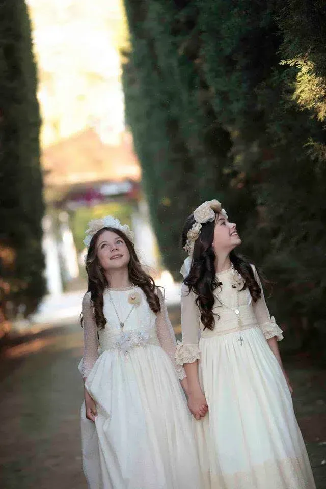 Dos niñas con vestidos blancos tomadas de la mano en un sendero arbolado al aire libre.