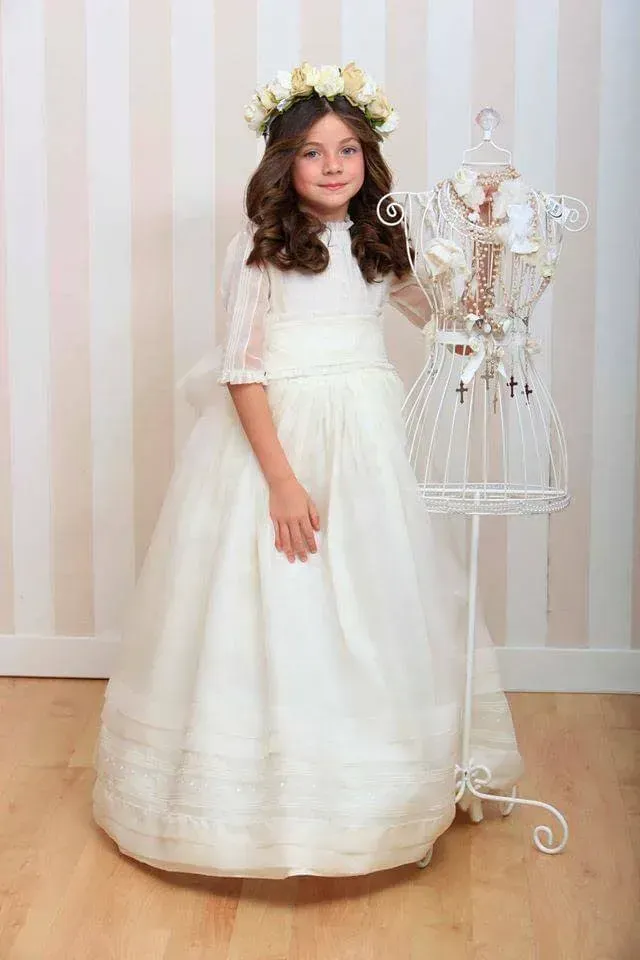 Chica con vestido blanco de pie junto a un maniquí sobre un fondo de papel tapiz a rayas.