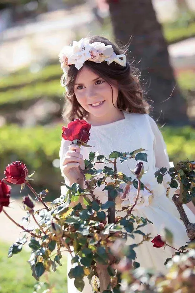 Niña sonriente con un vestido blanco sosteniendo una rosa roja en un jardín.