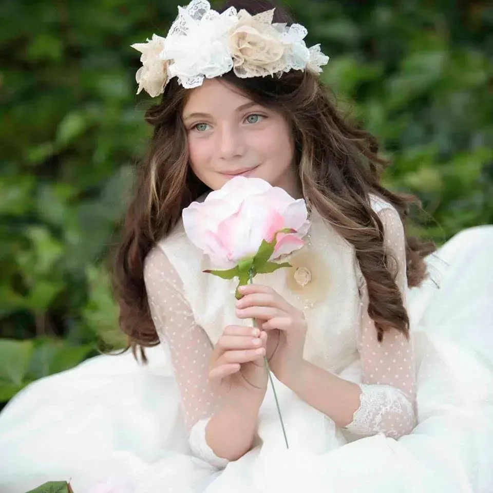 Mujer con vestido blanco sosteniendo una flor rosa, luciendo una corona floral al aire libre entre follaje verde.