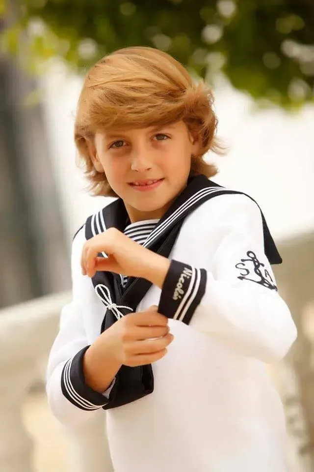 Niño con un traje blanco de estilo marinero sonriendo al aire libre con los brazos cruzados.