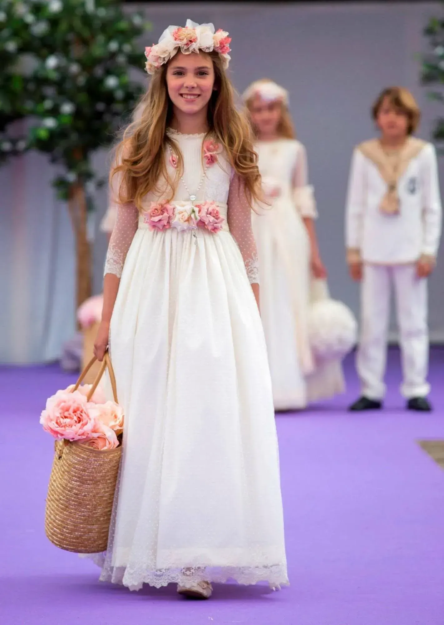 Una joven con un vestido blanco estampado de flores y una corona de flores lleva una cesta de mimbre en una pasarela morada.