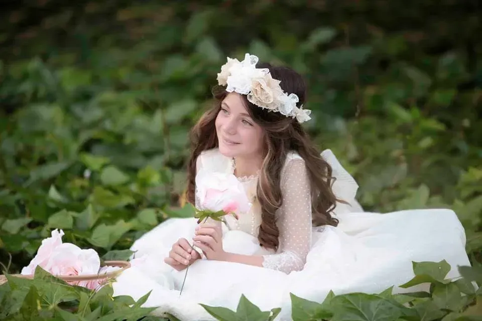 Mujer con vestido blanco y corona de flores, recostada sobre la hierba, sosteniendo un ramo de flores rosas.