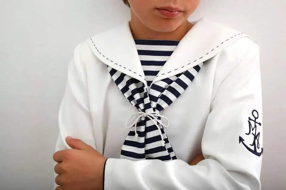 Niño con un traje de marinero azul marino y blanco con los brazos cruzados sobre un fondo liso.