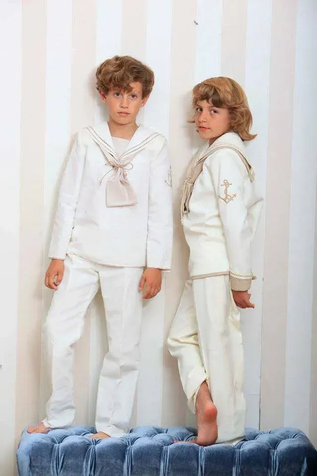Dos niños vestidos de blanco posando en un sofá azul con un fondo de rayas en tonos pastel.