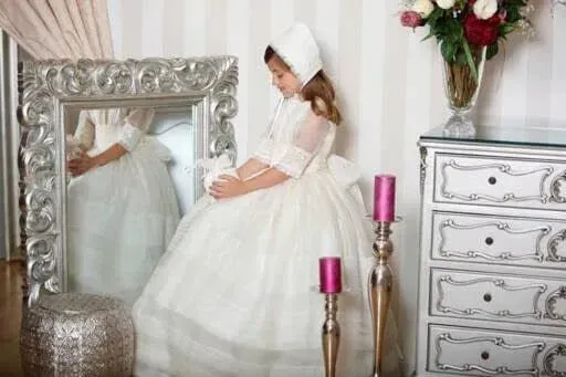 Niña con vestido blanco y gorro junto a un espejo y una cómoda ornamentados en una habitación luminosa.