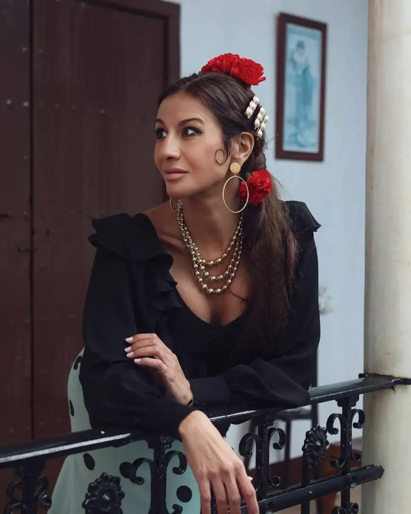 Mujer apoyada en la barandilla de un balcón, vestida con un vestido negro y perlas superpuestas, con flores rojas en el pelo.