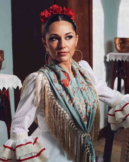 Mujer con blusa blanca bordada y chal con flecos, posando en interiores con un tocado de flores rojas.