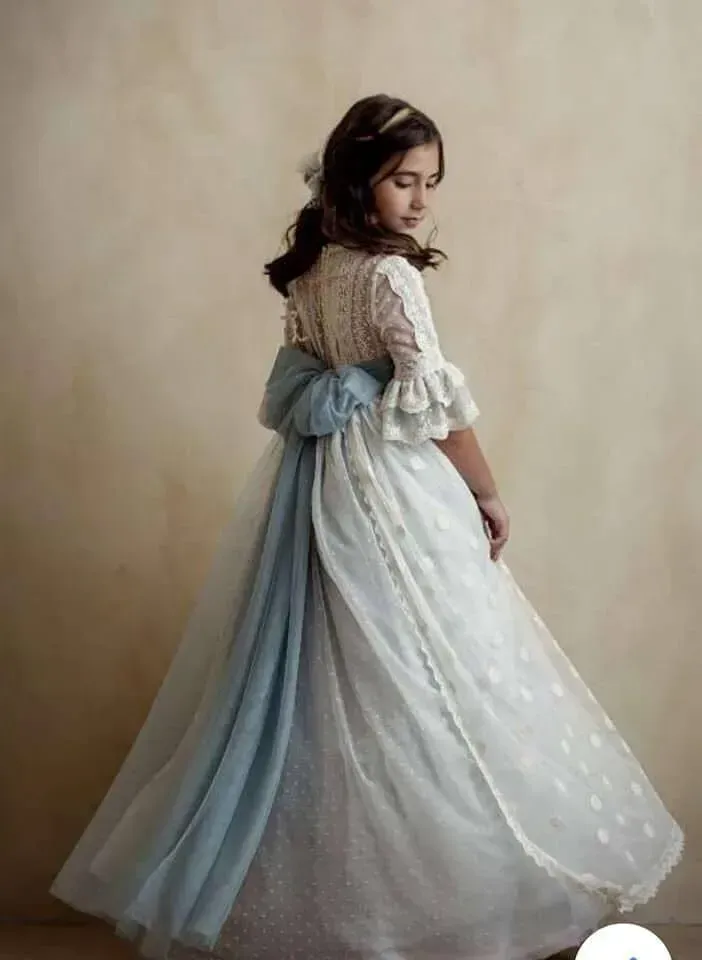Niña con un vestido blanco de encaje con una cinta azul, de pie y mirando por encima del hombro sobre un fondo beige.