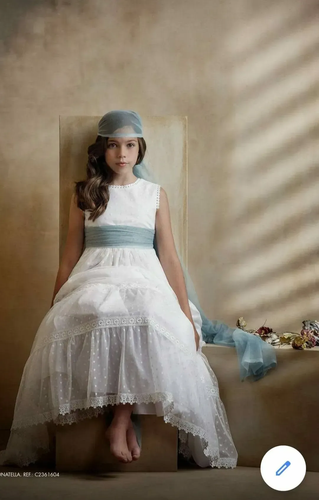 Una niña con un vestido blanco y un tocado azul está sentada junto a una pared beige con una suave luz a rayas.