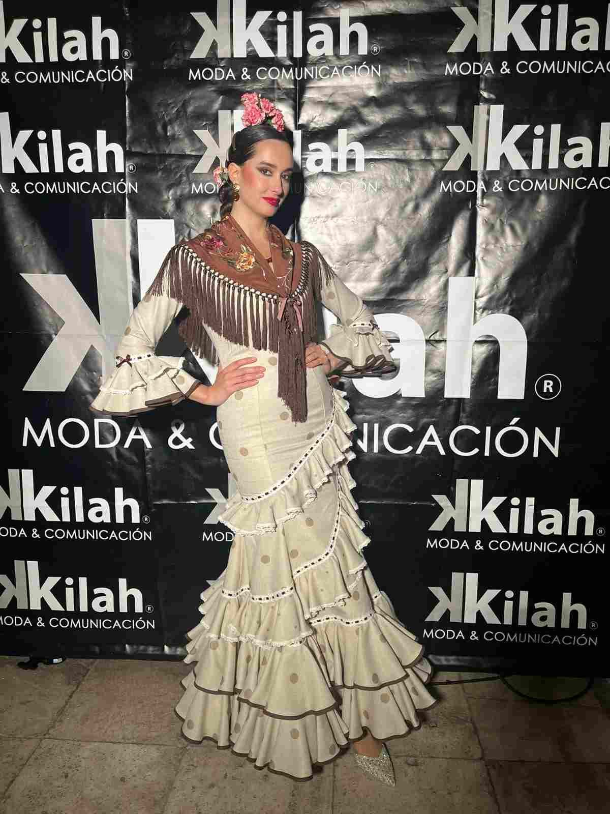 Mujer con un vestido plateado con volantes posando frente a un fondo de un evento de moda de Kilah