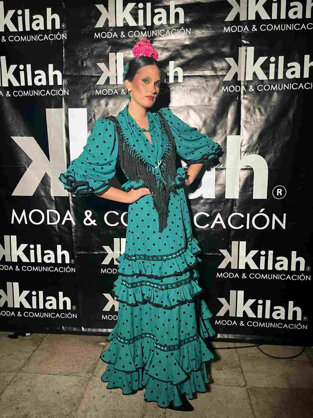 Mujer con un vestido de estilo flamenco color turquesa posando frente a un telón de fondo de Kilah Moda & Comunicación.