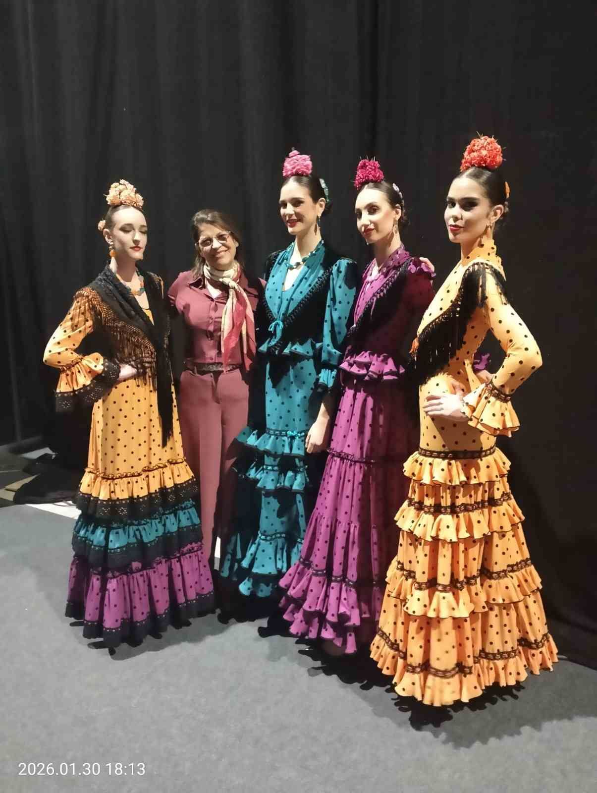Cinco bailarinas con coloridos trajes de flamenco posan en el escenario con un telón oscuro de fondo.