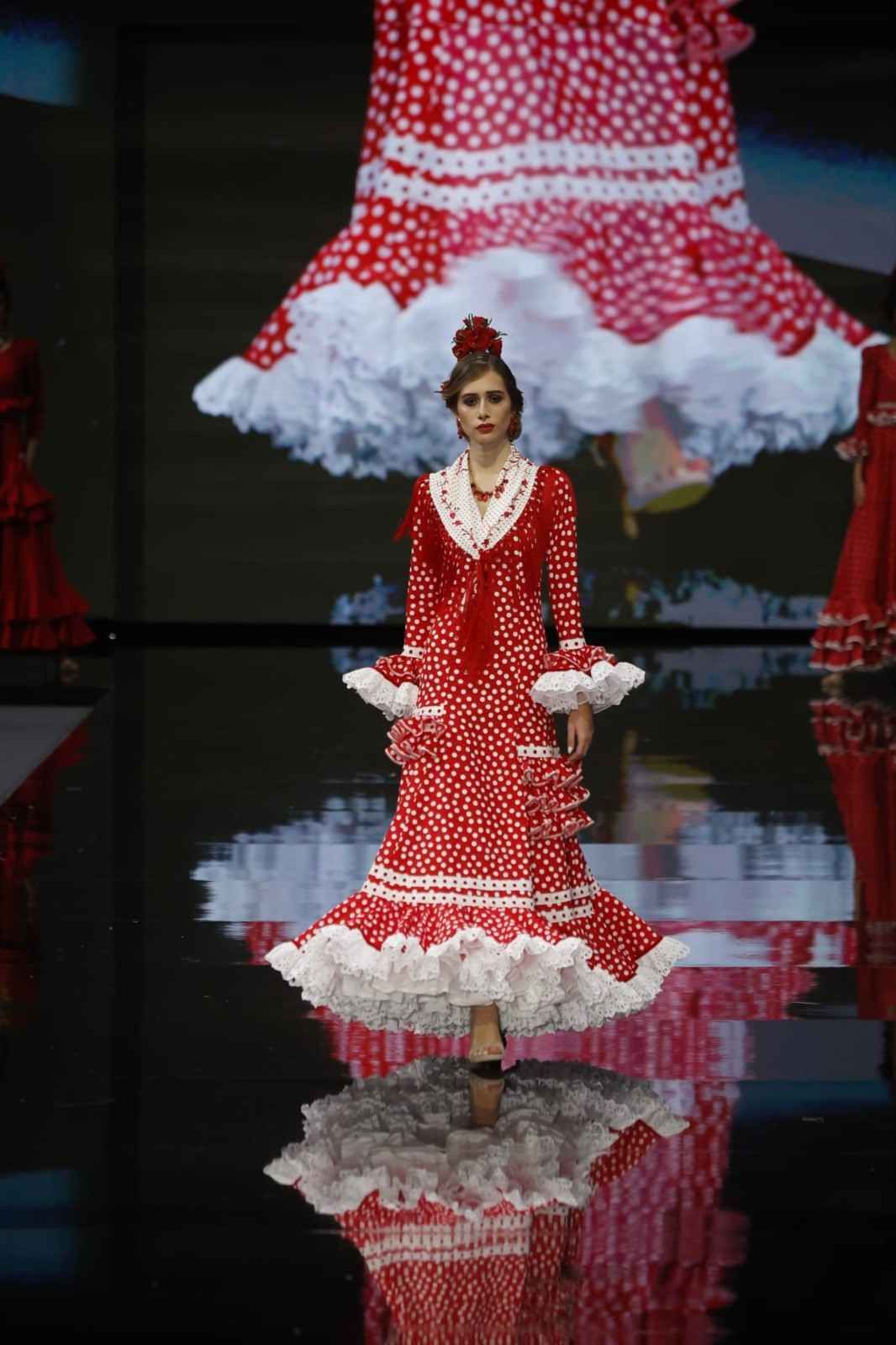 Una modelo con un vestido rojo de lunares y volantes blancos desfila por una pasarela, reflejada en un suelo negro brillante.