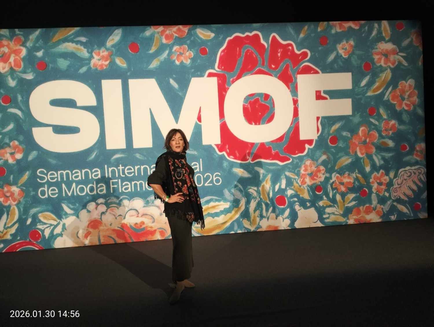 Persona de pie frente a un telón de fondo floral para eventos de SIMOF con texto blanco grande.