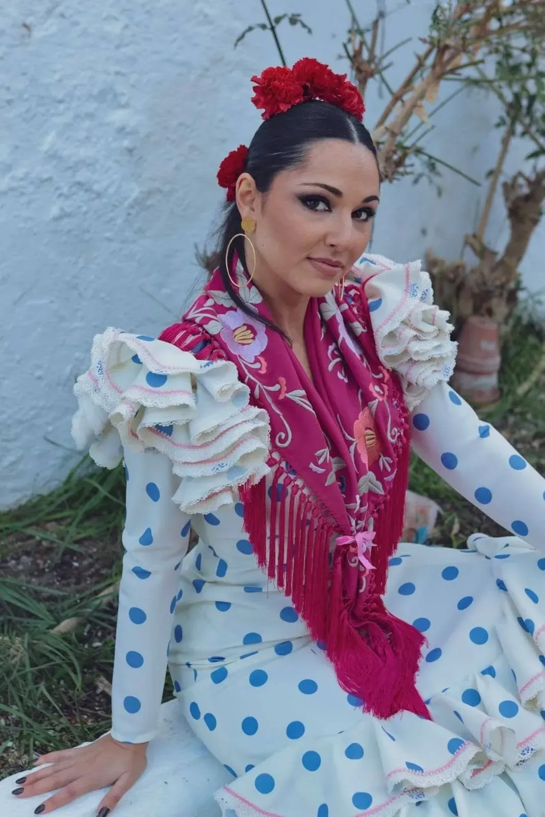 Mujer con un vestido blanco de lunares azules y un chal rojo, sentada al aire libre contra una pared de color claro.