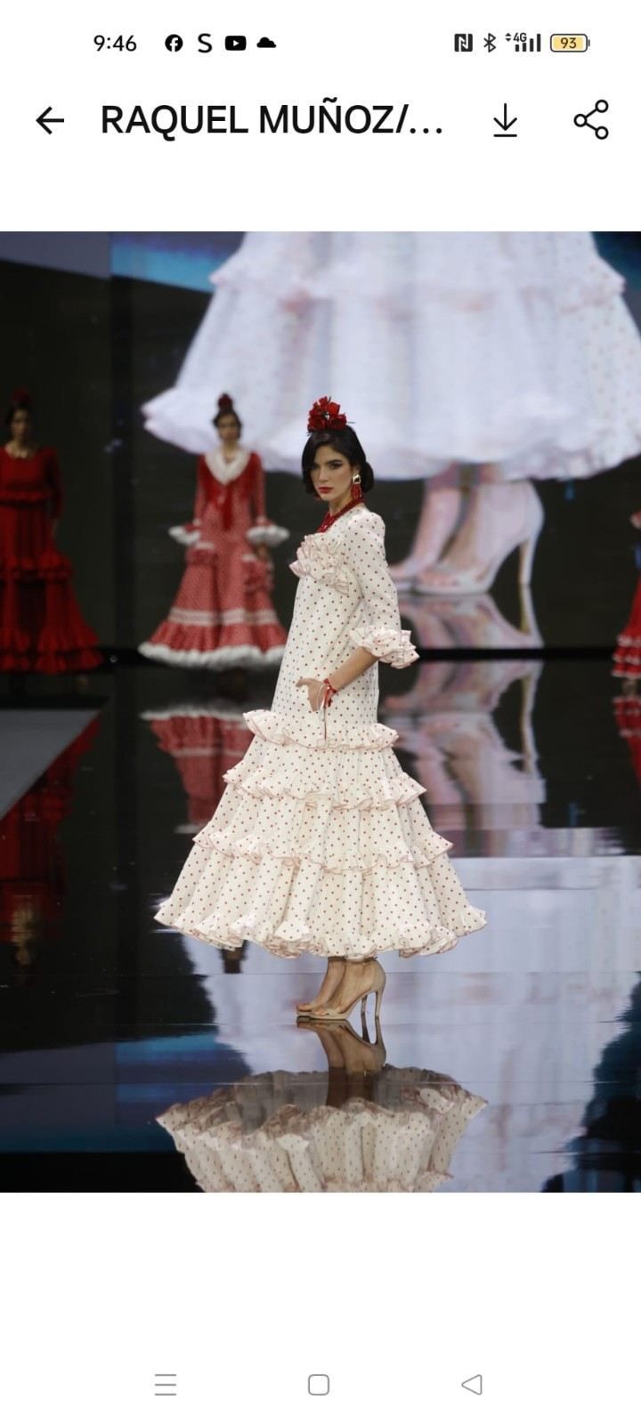 Una modelo desfila con un vestido color crema con volantes y un sombrero sobre una pasarela reflectante.