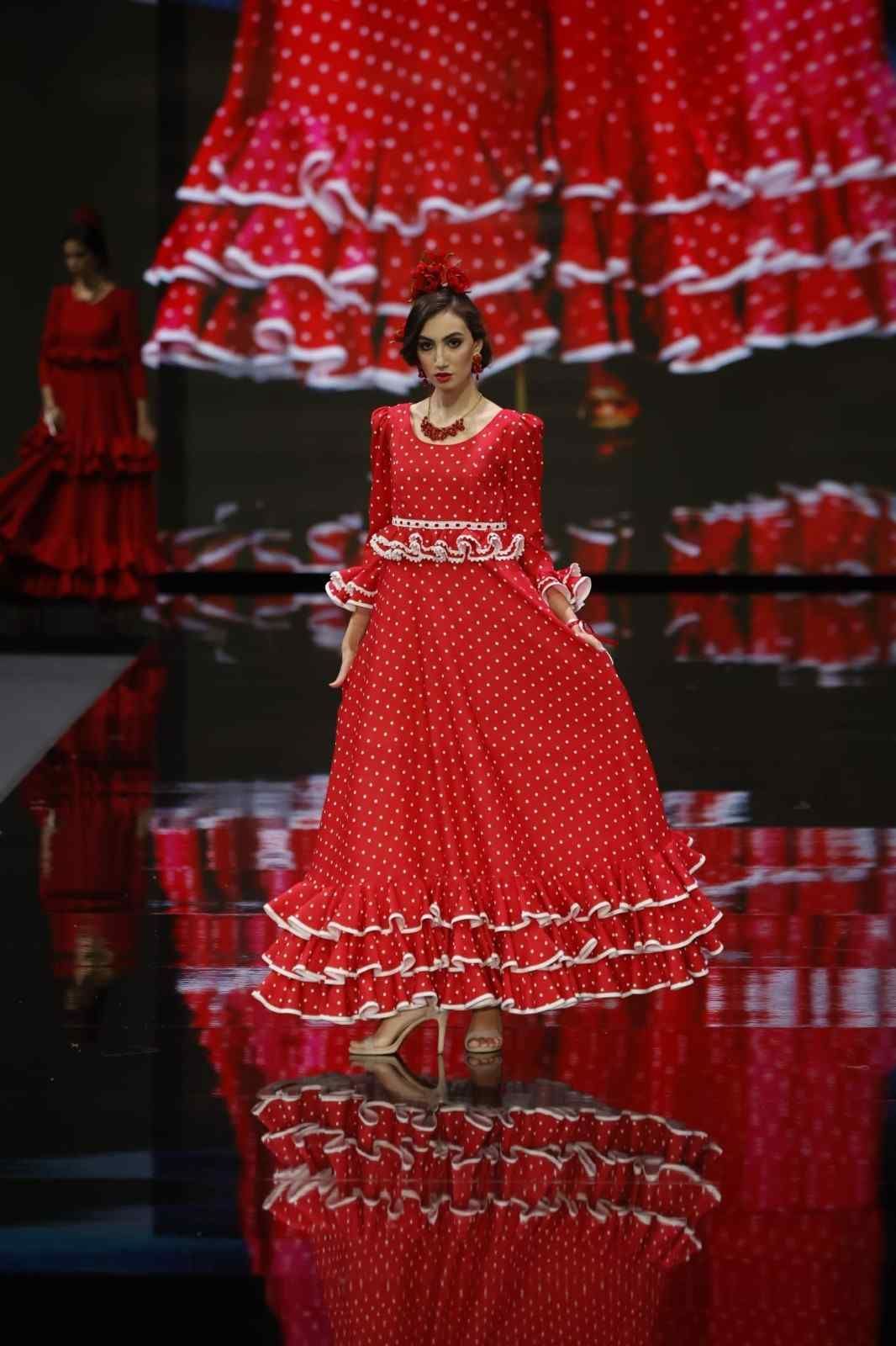 Modelo con un vestido rojo con volantes desfilando por una pasarela con suelo negro reflectante y fondo rojo.