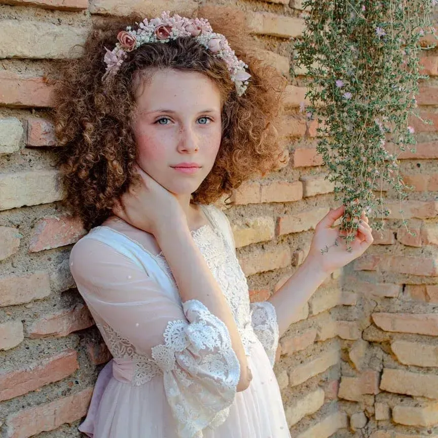Mujer con vestido blanco de encaje y corona de flores posando junto a una pared de ladrillos.