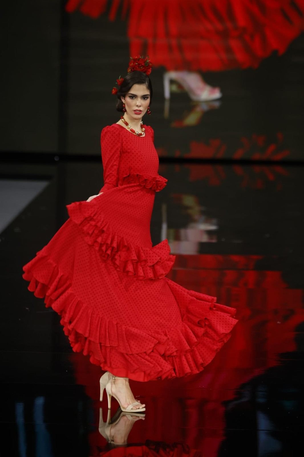 Una modelo con un vaporoso vestido rojo con volantes desfila por una pasarela bajo una iluminación espectacular.