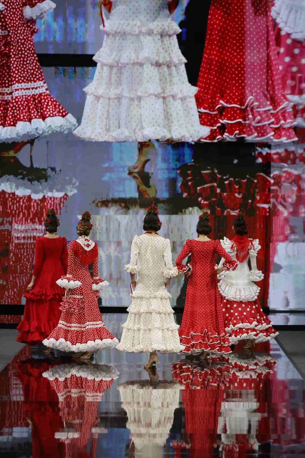 Modelos con vestidos rojos y blancos con volantes desfilan en una pasarela con un suelo negro reflectante.