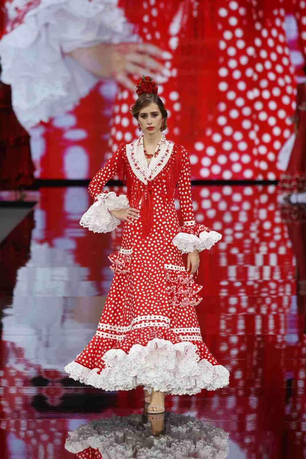 Modelo con vestido de lunares rojos y blancos desfilando por una pasarela con fondo rojo.