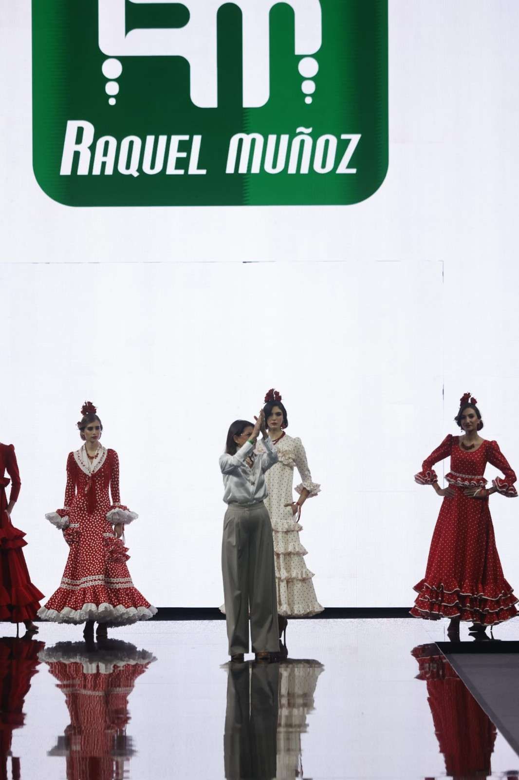 Cuatro bailarinas con vestidos de estilo flamenco rojos y grises en un escenario con un telón de fondo de Raquel Muñoz.