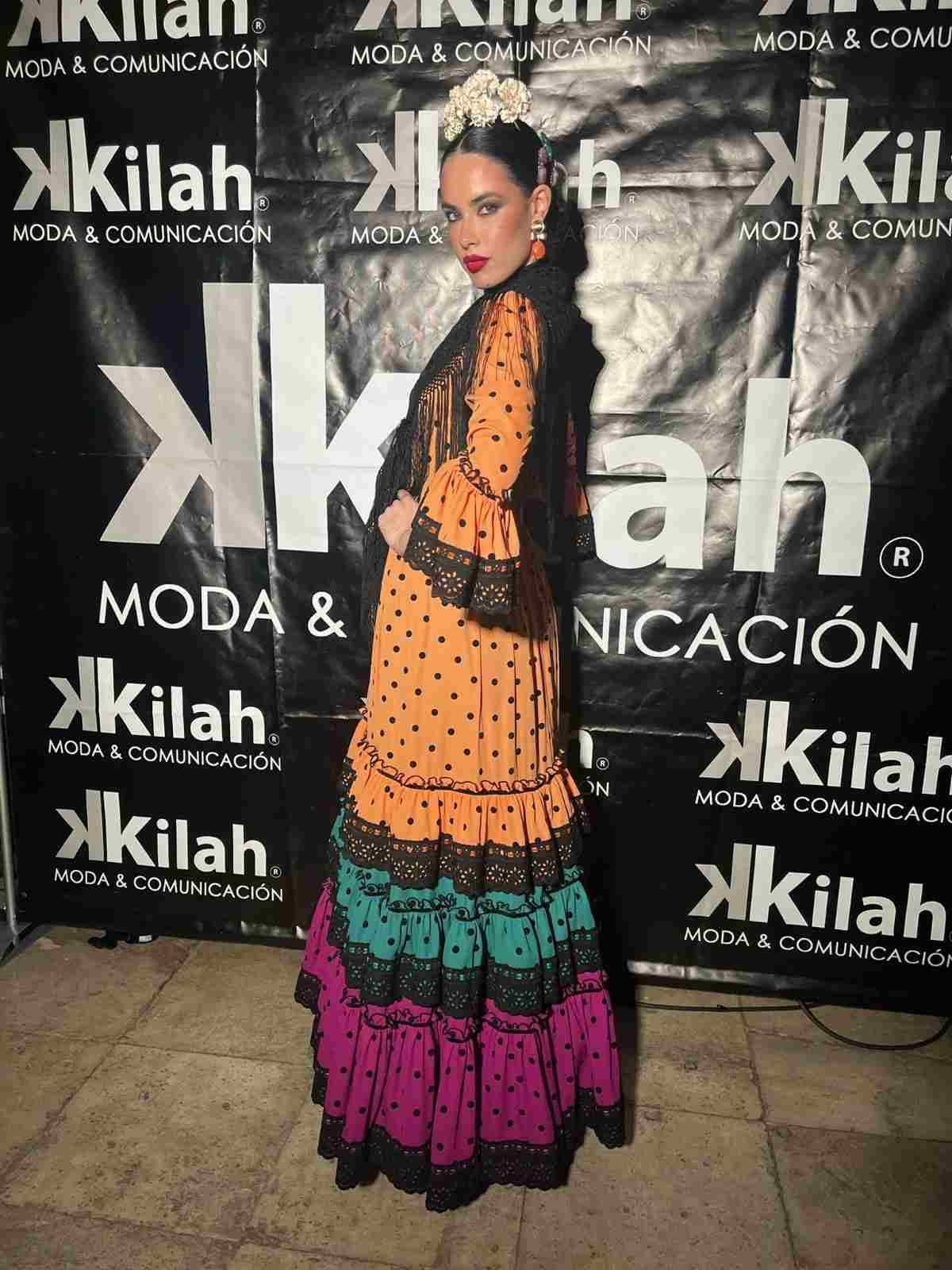 Mujer posando sobre un fondo estampado, luciendo un vestido de lunares negros y naranjas con volantes rosas y verdes.