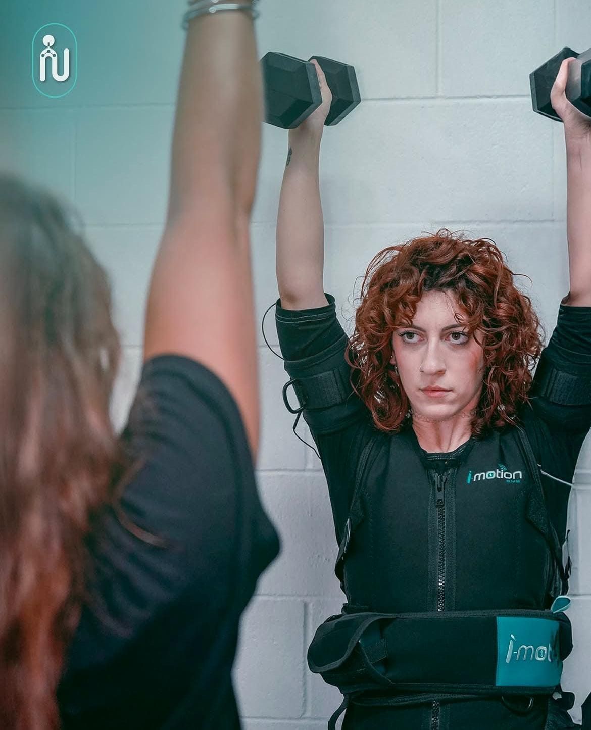 Mujer levantando mancuernas en un gimnasio, vista reflejada en un espejo, con expresión concentrada.