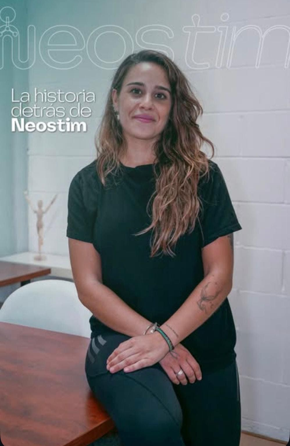 Mujer sentada en un escritorio, vestida de negro, sonriendo frente a un póster de Neostim.