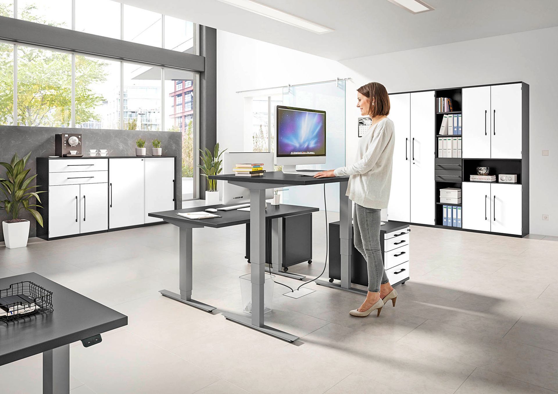Frau steht an einem höhenverstellbaren Schreibtisch, dieser steht in einem schlicht eingerichtetem Büro