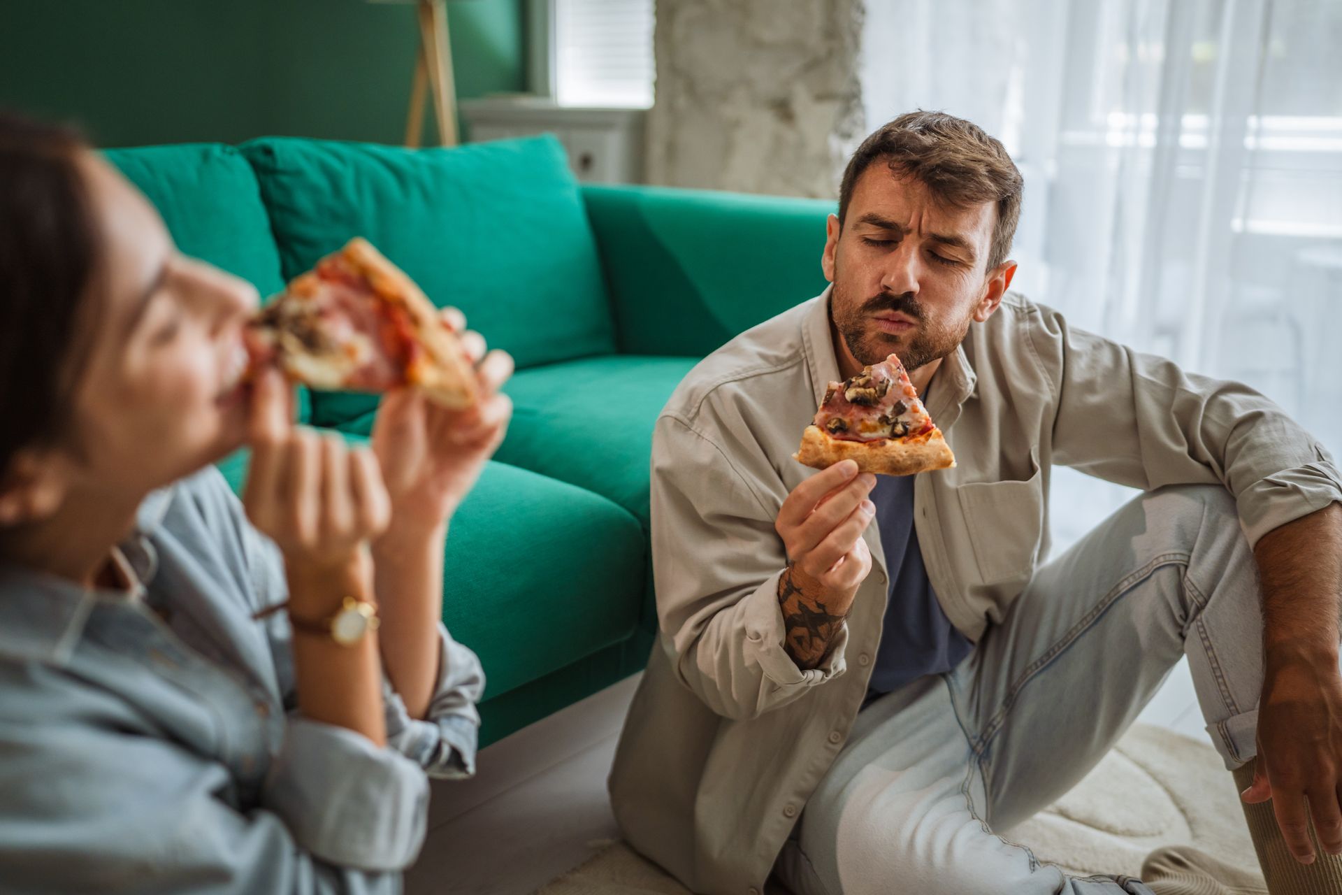 Deux personnes mangent une pizza dans un salon lumineux, avec un canapé turquoise en arrière-plan