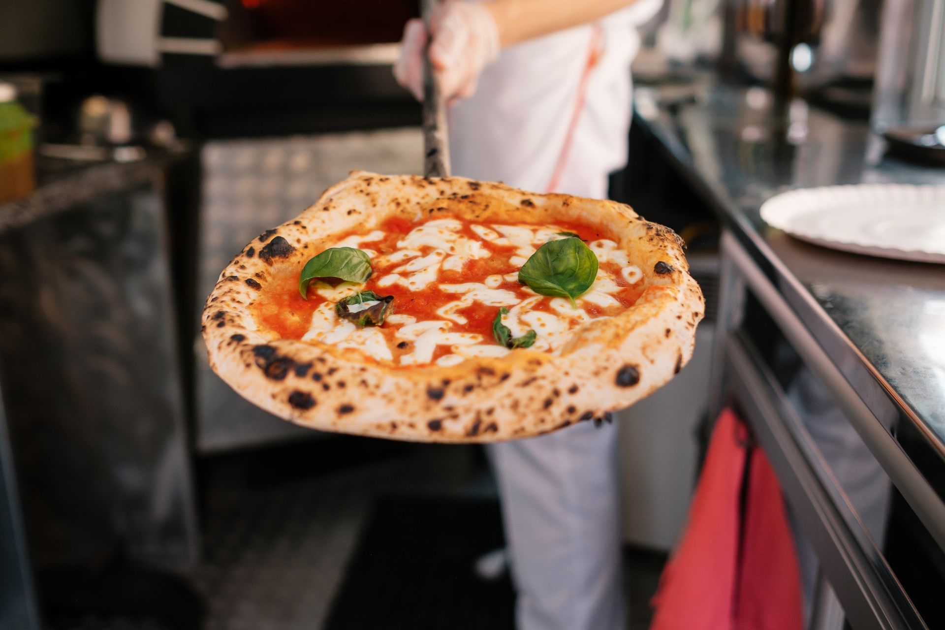 Une pizza fraîche est présentée dans la cuisine d'une pizzeria, avec de la sauce tomate et des feuilles de basilic sur la pâte