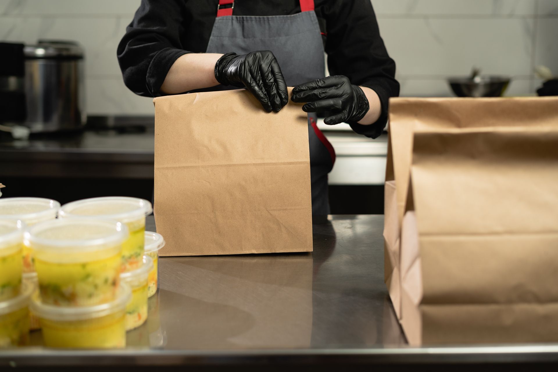 Des mains gantées remplissent des sacs à repas en papier brun sur le comptoir de la cuisine, avec des contenants alimentaires à proximité