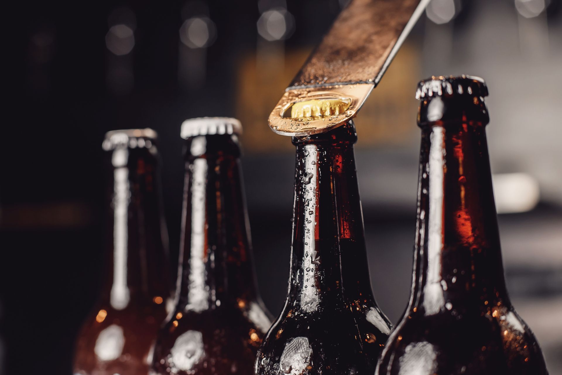 Des bouteilles de bière, un ouvre-bouteille en train d'enlever une capsule, posées sur un comptoir de bar sombre