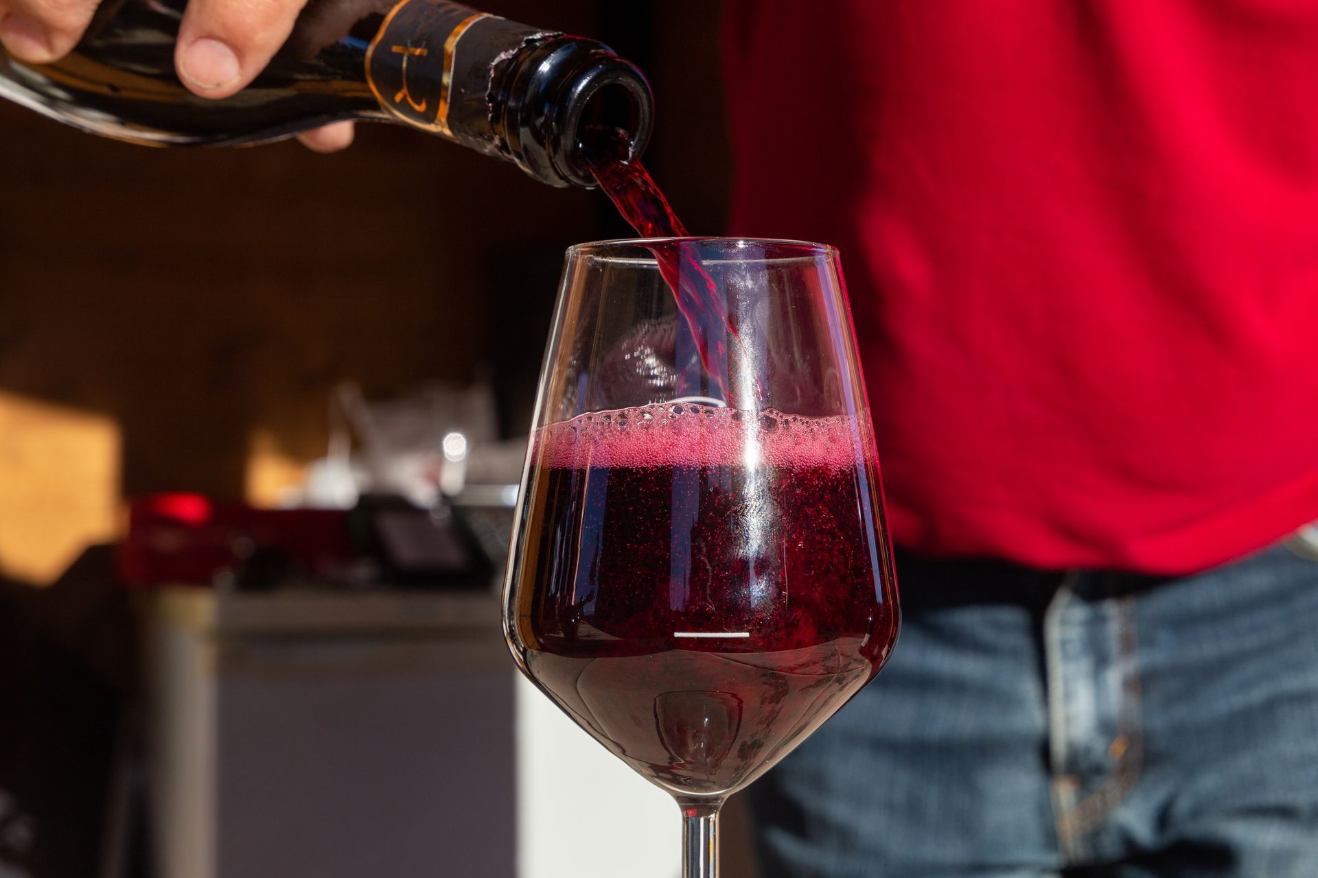 Du vin est versé dans un verre à une table, avec une personne en chemise rouge en arrière-plan