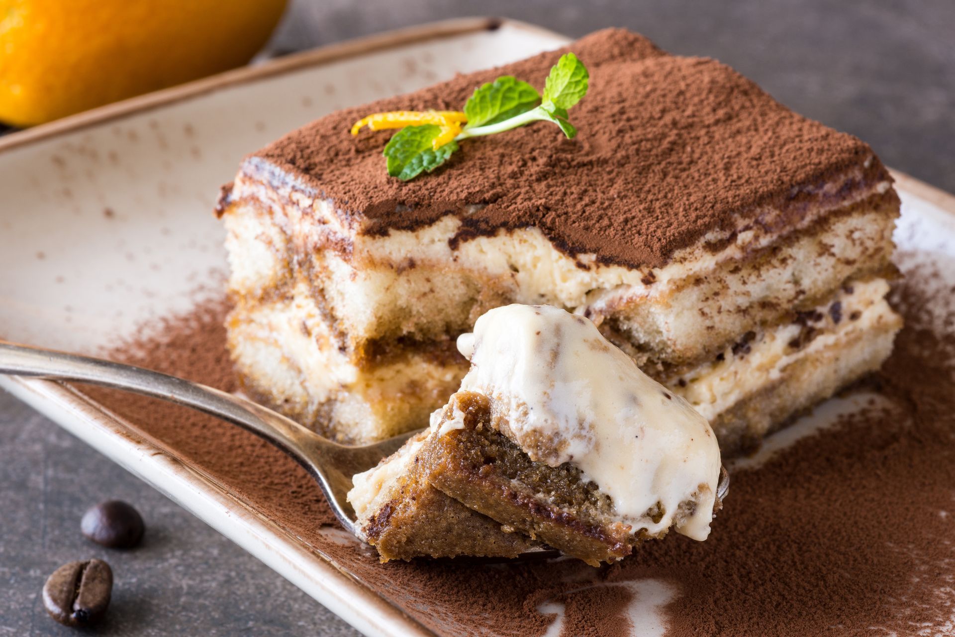 Une part de tiramisu sur une assiette, saupoudrée de cacao et garnie de menthe, avec une cuillère à côté