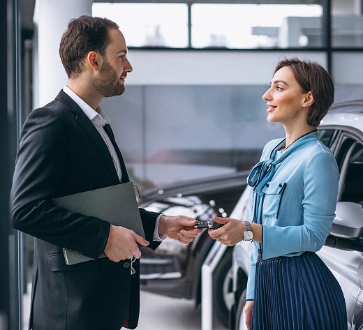 Dos personas conversando e intercambiando llaves en una sala de exposición de automóviles, con un vehículo al fondo.