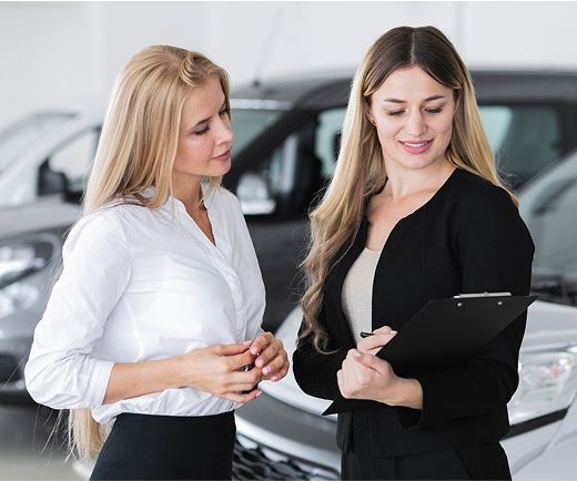 Dos mujeres conversando sobre un coche en una sala de exposición; una de ellas sostiene un portapapeles.