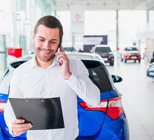 Hombre sonriente con camisa blanca hablando por teléfono y sosteniendo un portapapeles junto a un coche azul