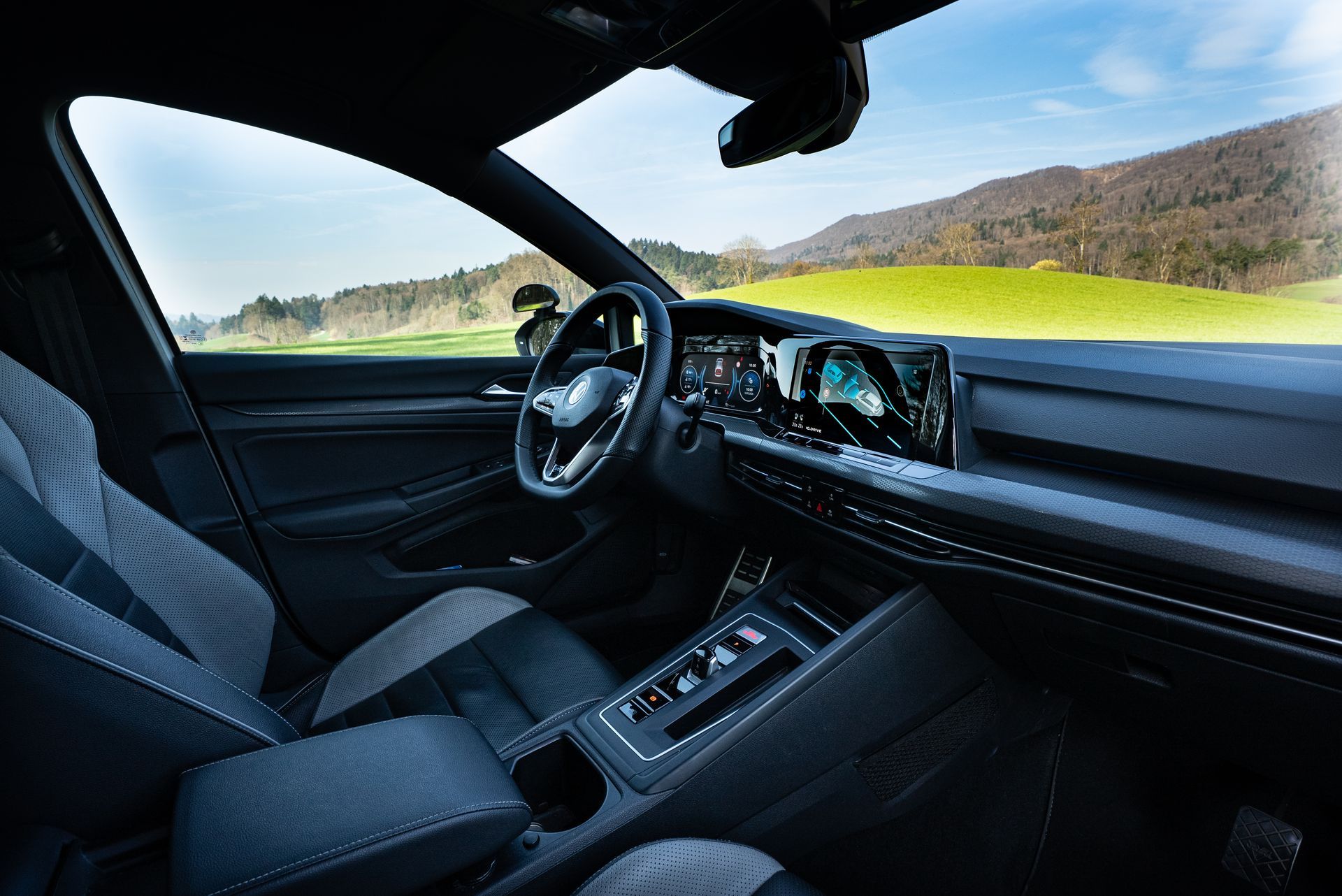 Eine Person stellt orangefarbene Verkehrskegel auf einem Asphaltparkplatz auf; im Hintergrund ist ein Auto zu sehen, helles Sonnenlicht.