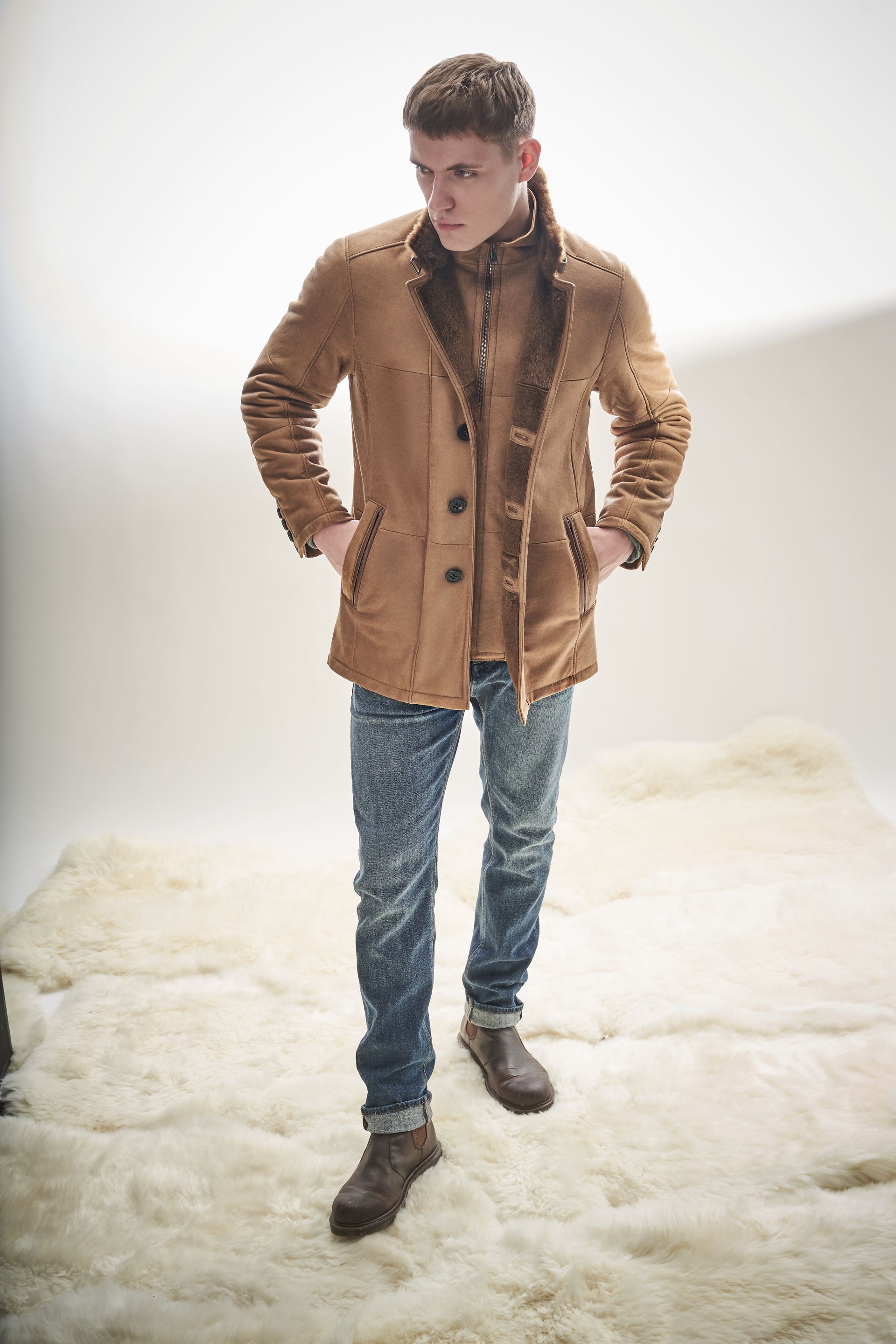 Ein Mann in einem braunen Mantel und Jeans steht auf einem weißen Teppich.