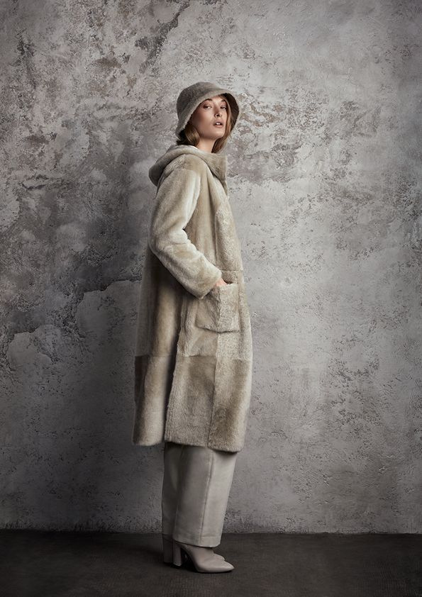 Eine Frau mit Pelzmantel und Hut steht vor einer Betonmauer.