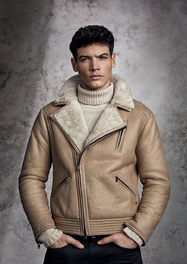 Ein Mann in einer hellbraunen Jacke und einem weißen Rollkragenpullover steht mit den Händen in den Taschen da.