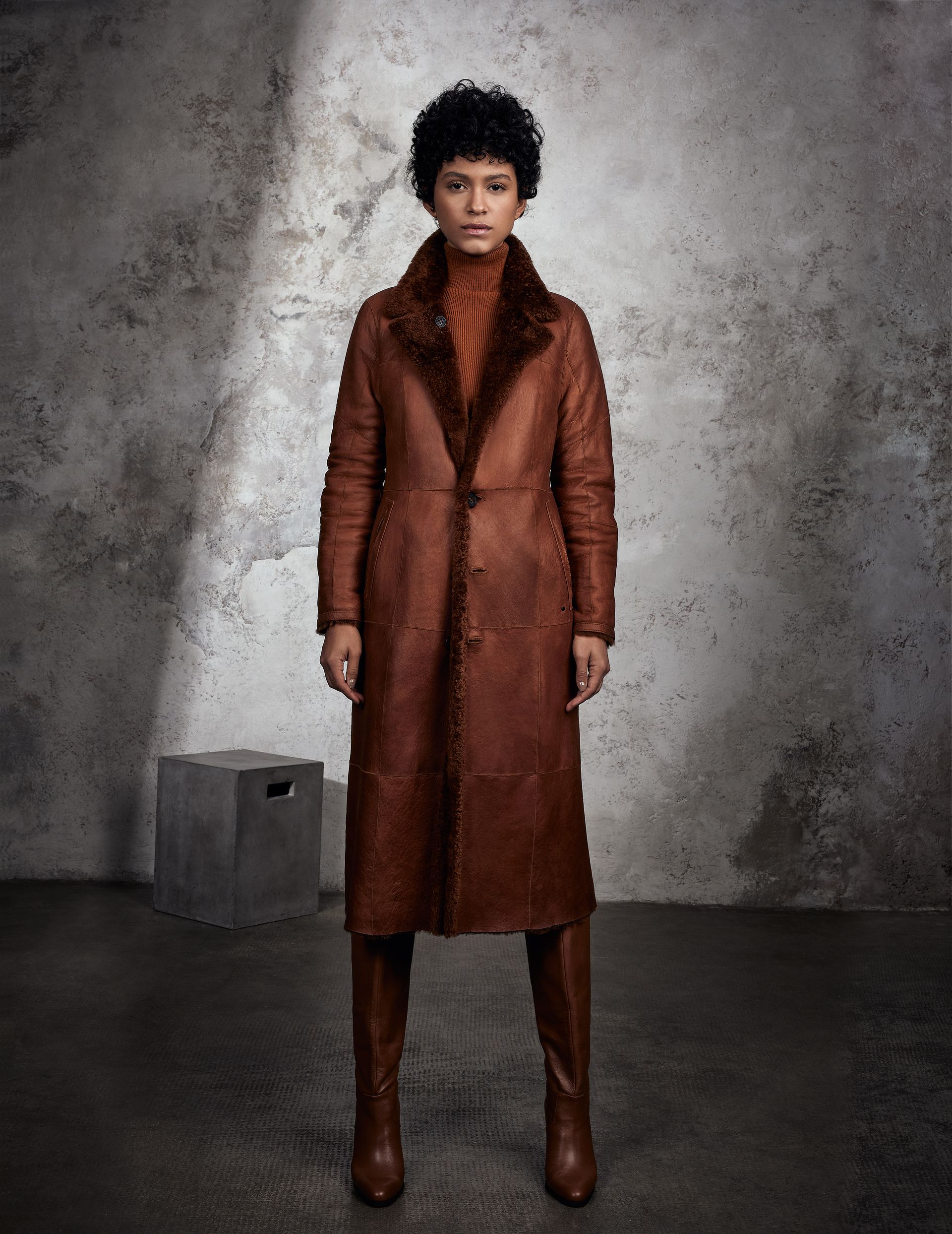 Eine Frau mit einem braunen Mantel und Stiefeln steht vor einer Wand.