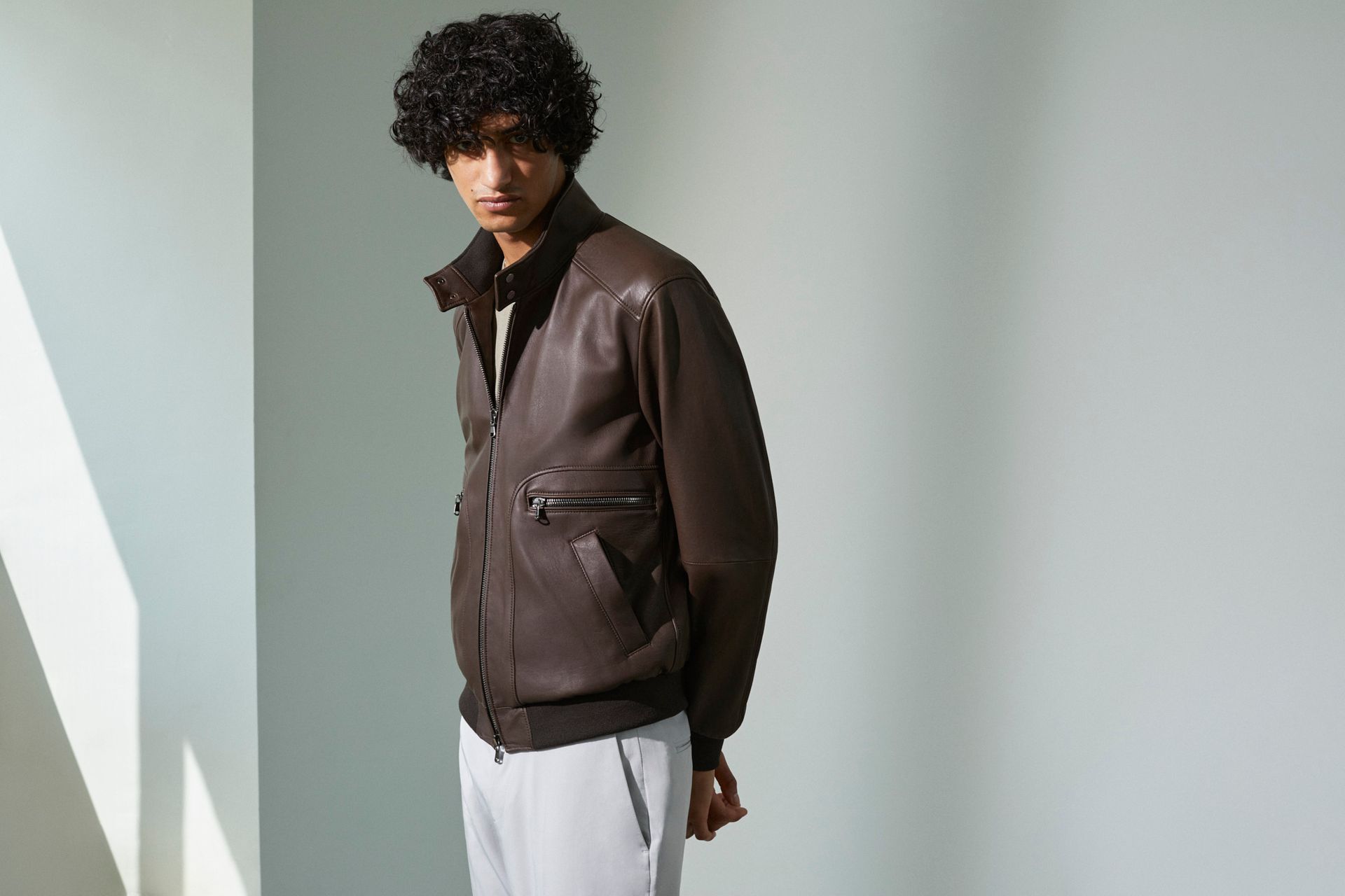 Ein Mann mit einer braunen Lederjacke und weißen Hosen steht in einem Raum.
