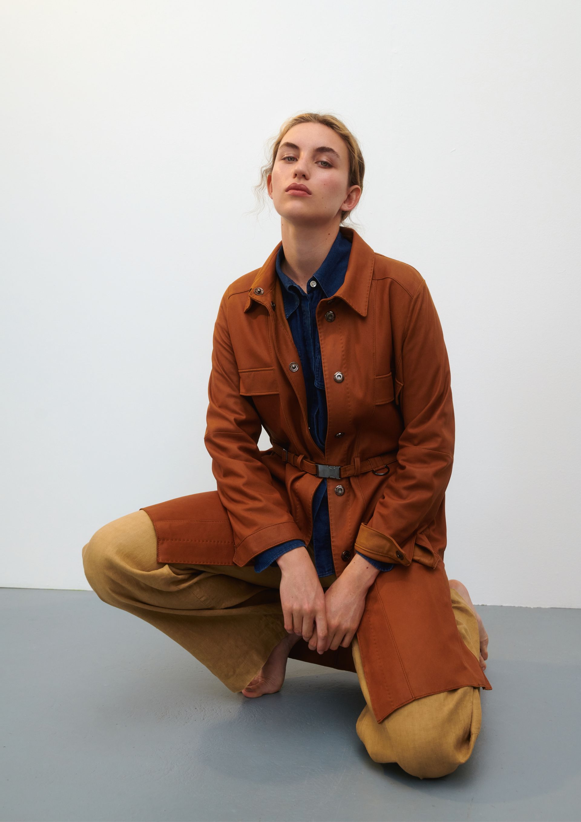 Eine Frau kniet nieder und trägt eine braune Jacke und hellbraune Hosen.