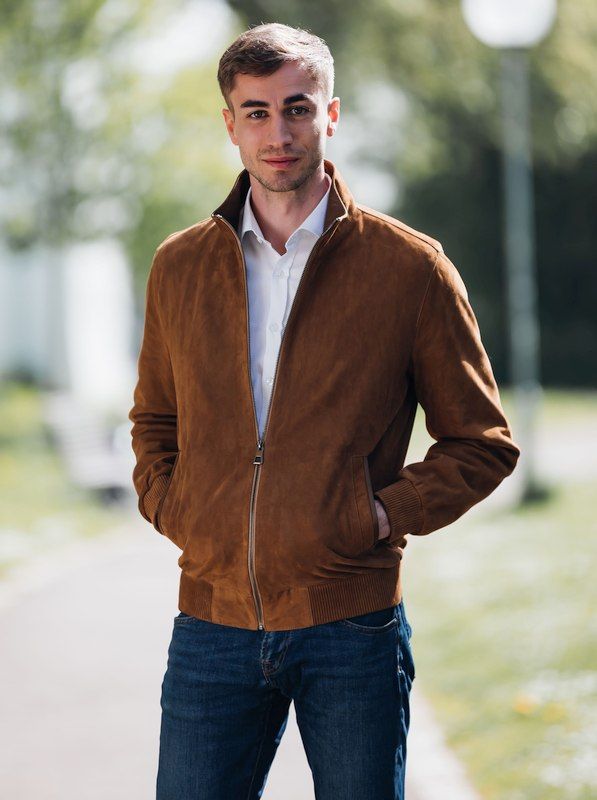 Ein junger Mann in einer braunen Wildlederjacke und Jeans steht auf einem Bürgersteig.